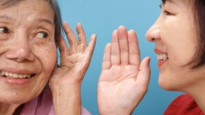 An elderly woman with gray hair and a smile cups her ear while listening to a younger woman, who is smiling and speaking close to her ear. The background is a solid blue color.