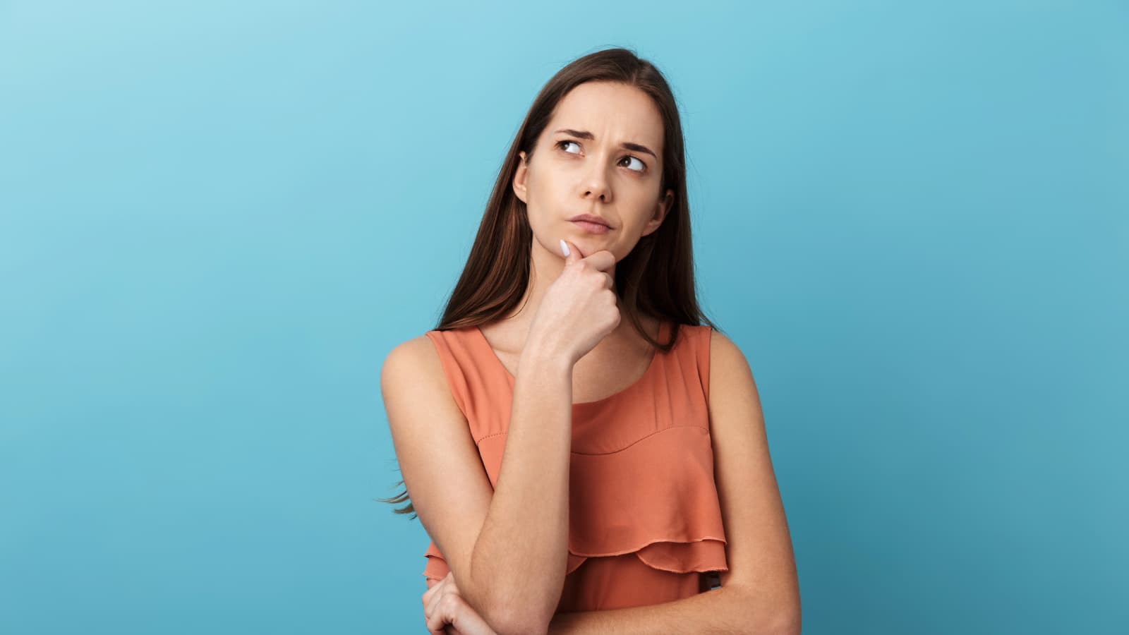 A young woman with long brown hair wearing an orange sleeveless top, standing against a solid blue background, looking up with a thoughtful expression, resting her chin on her hand.